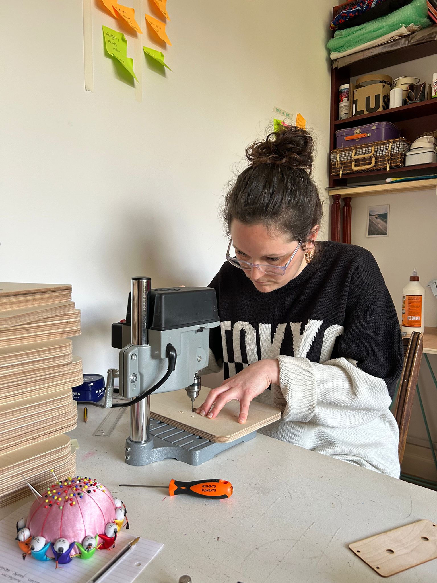 Artist Rianne Houba is sitting behind a desk and using a small pillar drill to drill holes into pieces of plywood.
