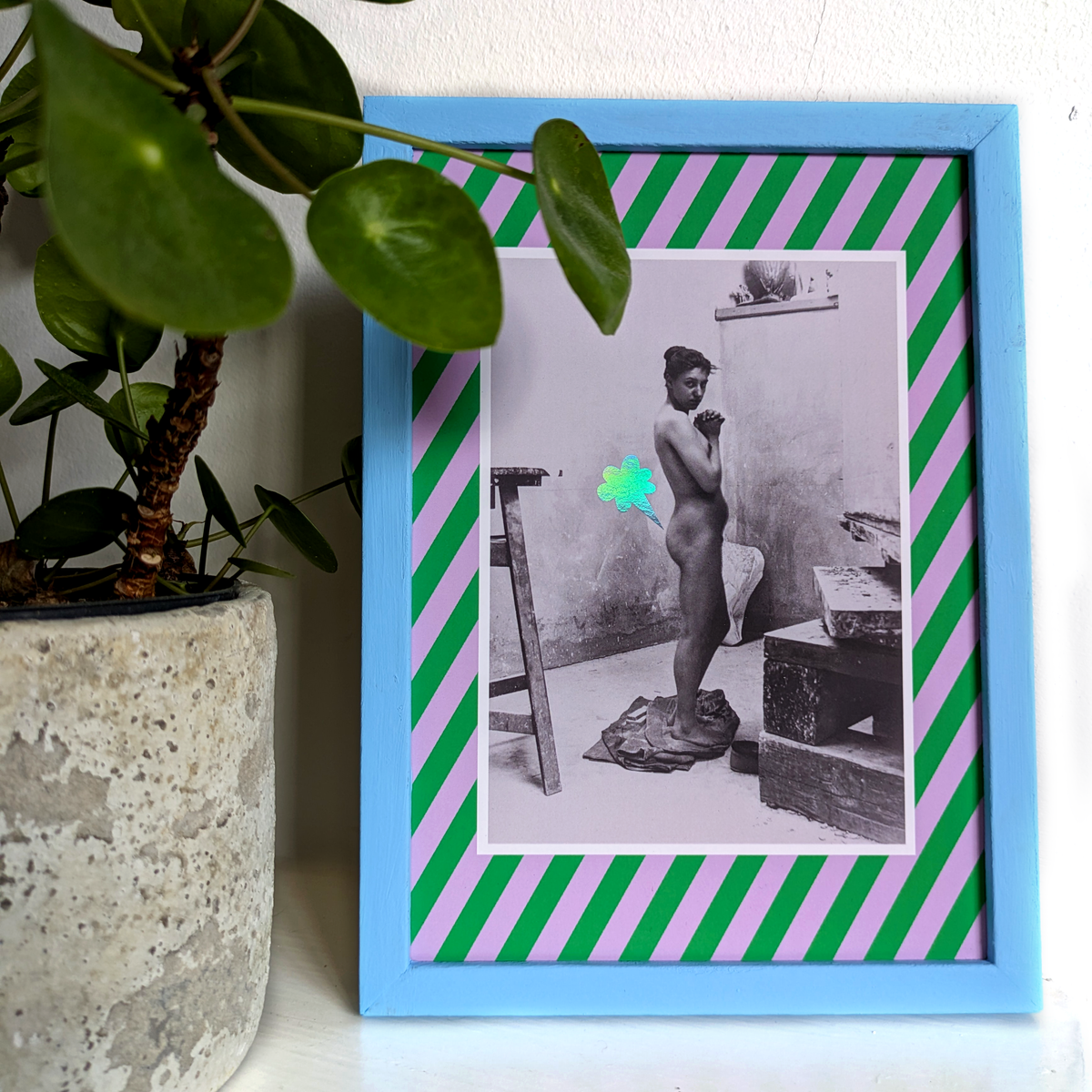 A playful poster featuring a photograph of a nude female standing in an art studio framed by a green and lilac striped border. A cartoon-style fart cloud, printed in holographic foil, is placed next to her bum. The poster is shown inside a blue frame and is standing next to a plant.
