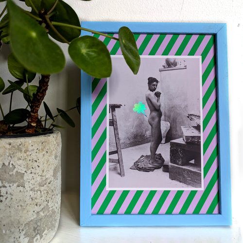 A playful poster featuring a photograph of a nude female standing in an art studio framed by a green and lilac striped border. A cartoon-style fart cloud, printed in holographic foil, is placed next to her bum. The poster is shown inside a blue frame and is standing next to a plant.