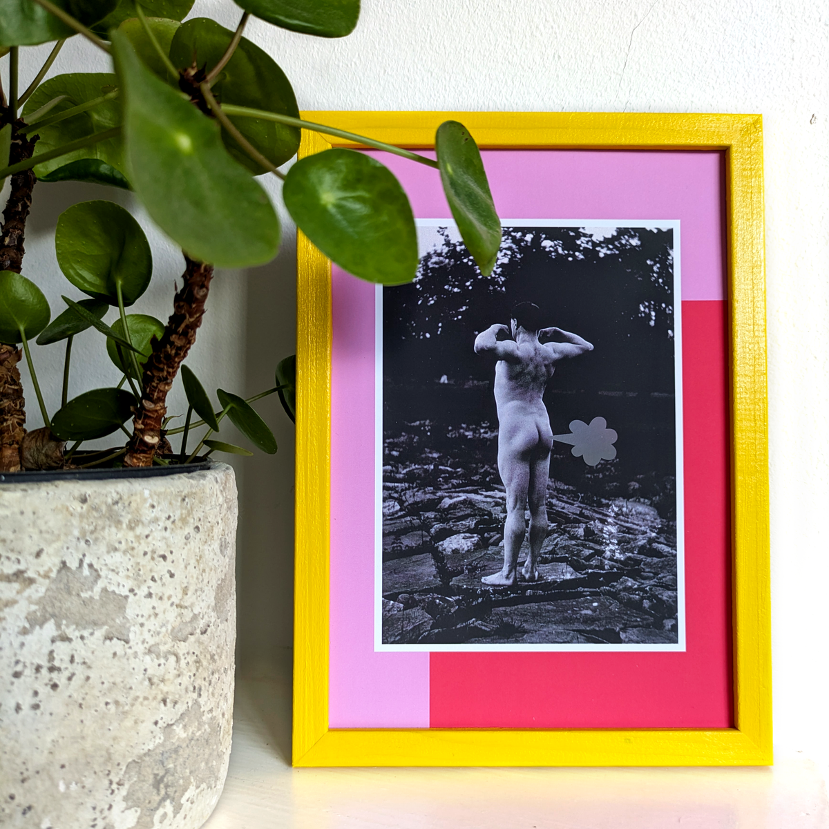 A playful poster featuring a photograph of a nude male flexing outdoors framed by a red and pink graphic border. A cartoon-style fart cloud, printed in holographic foil, is placed next to his bum. The poster is shown inside a yellow frame and is standing next to a plant.
