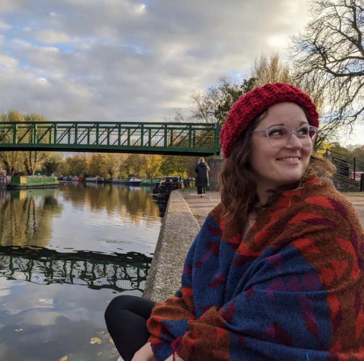 Artist Rianne Houba wearing a red beanie and colourful scarf sitting by a canal with a bridge in the background.