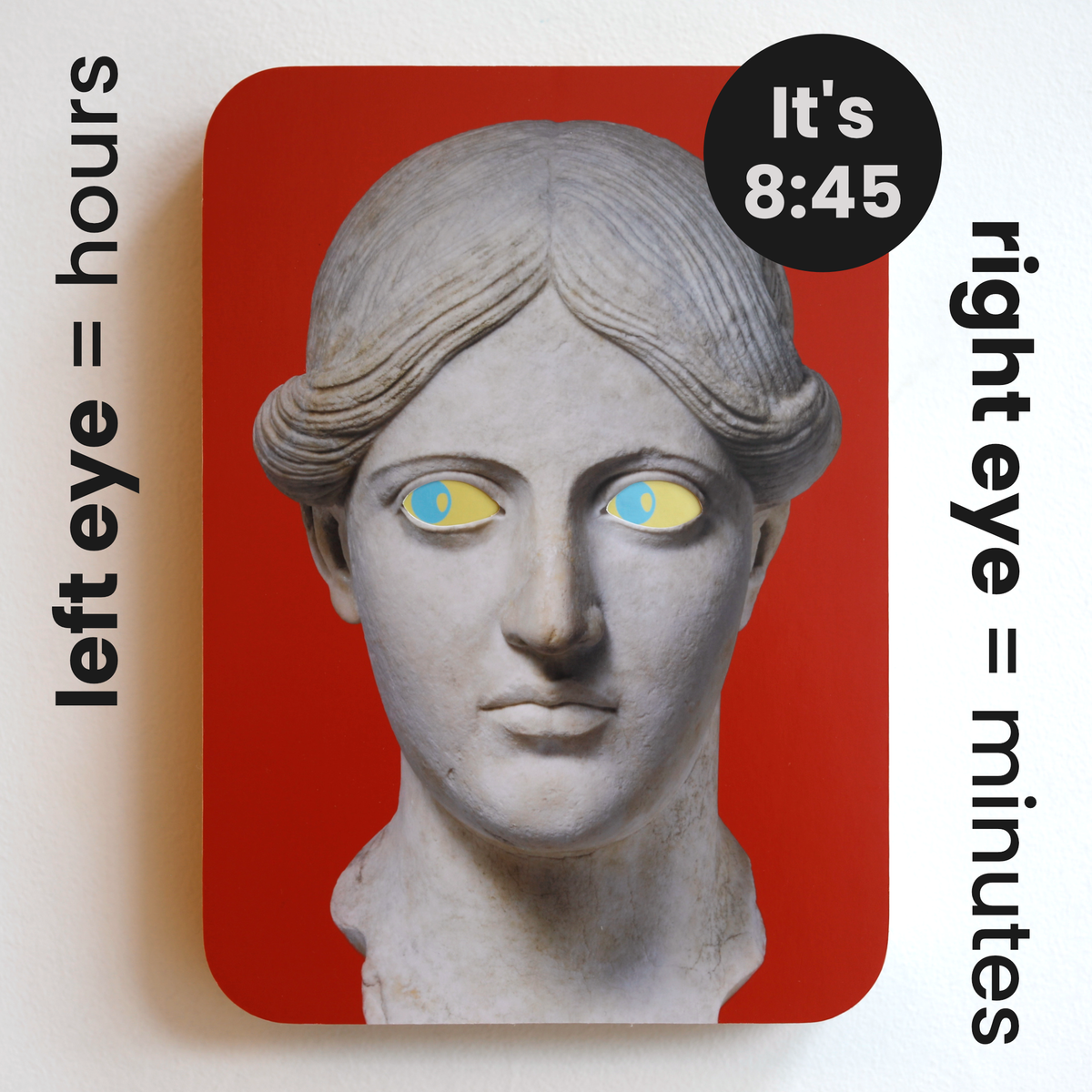 A printed statue head on a red rectangular background with yellow eyes and blue pupils that both look to the left. Image text: ‘Left eye = hours. Right eye = minutes. It’s 8:45.’