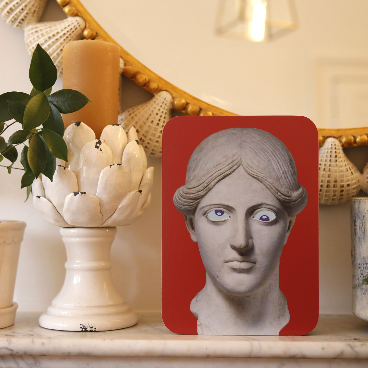 A unique art clock made of a printed statue head on a red rectangular background. The art piece is placed on a marble fireplace in front of a large mirror.