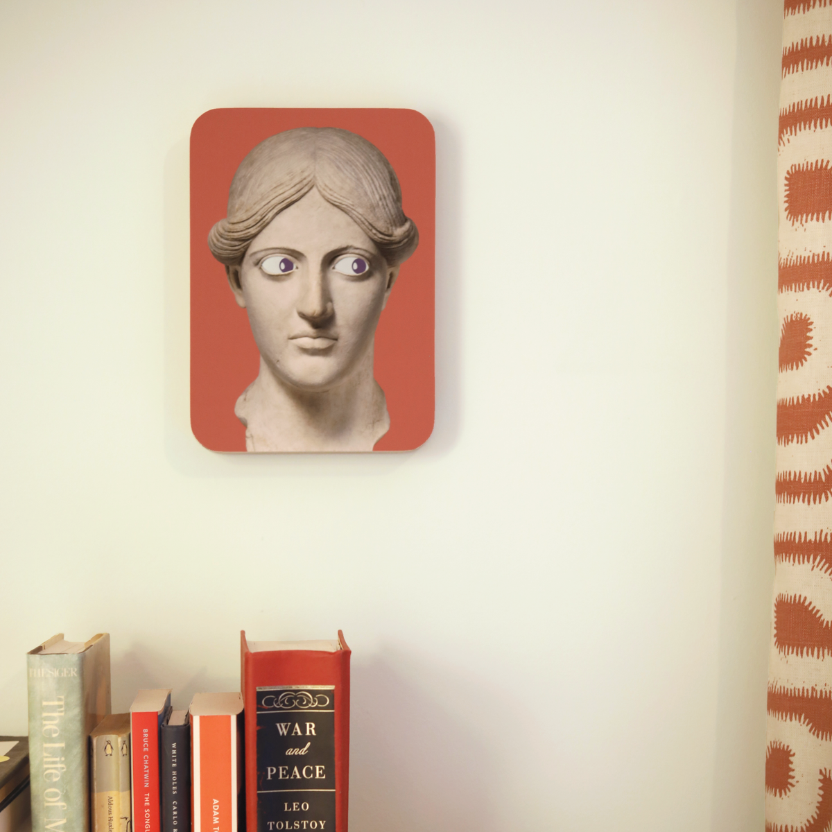 A unique art clock made of a printed statue head on a pink rectangular background hanging above a row of books and next to a curtain.