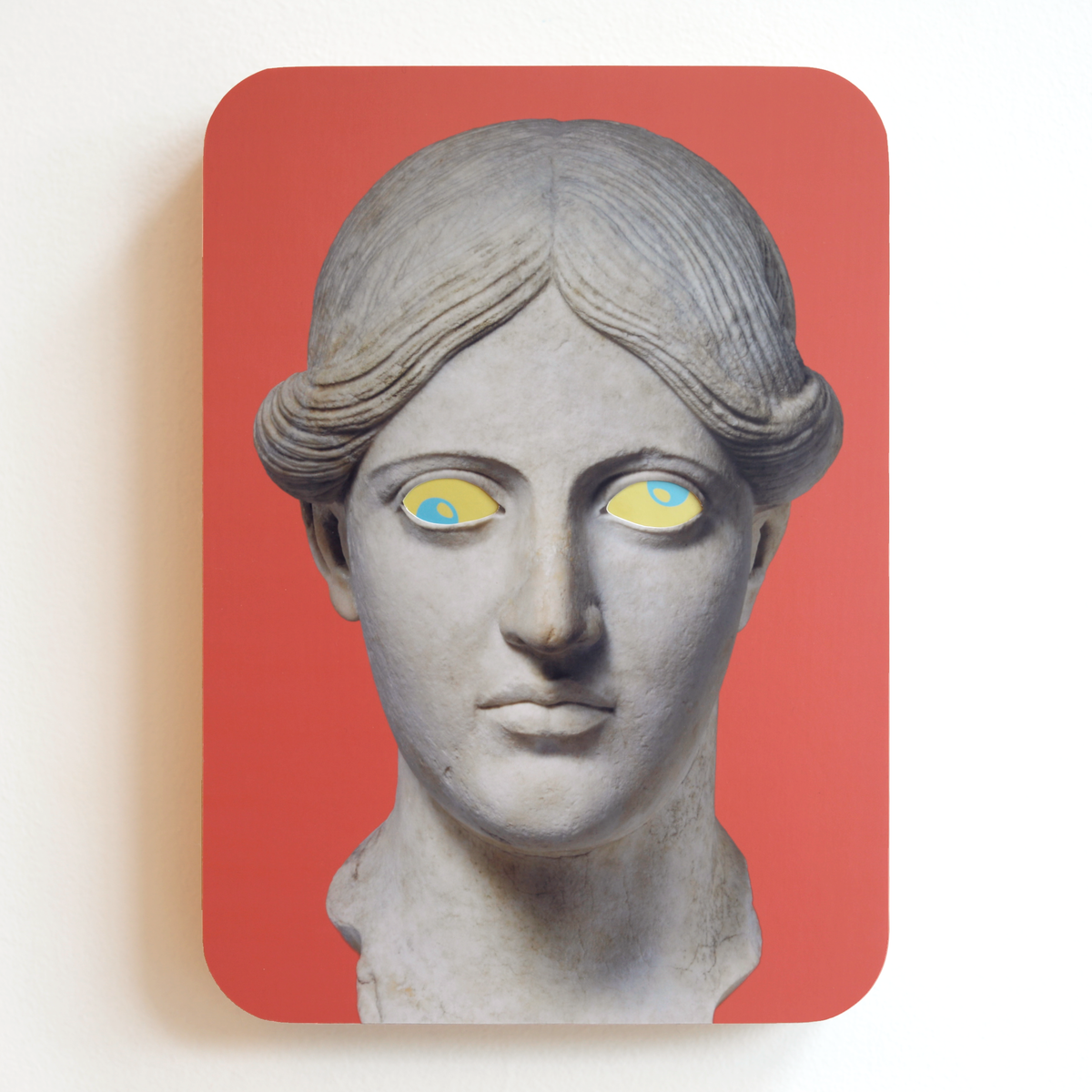 A printed statue head on a pink rectangular background with yellow eyes and blue pupils. One eye is looking up, the other eye is looking down.