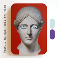 A unique art clock made of a printed statue head with blue and purple eyes on a red rectangular background. One eye is looking up, the other is looking down. Image text: ‘Psst... my eyes tell the time'.